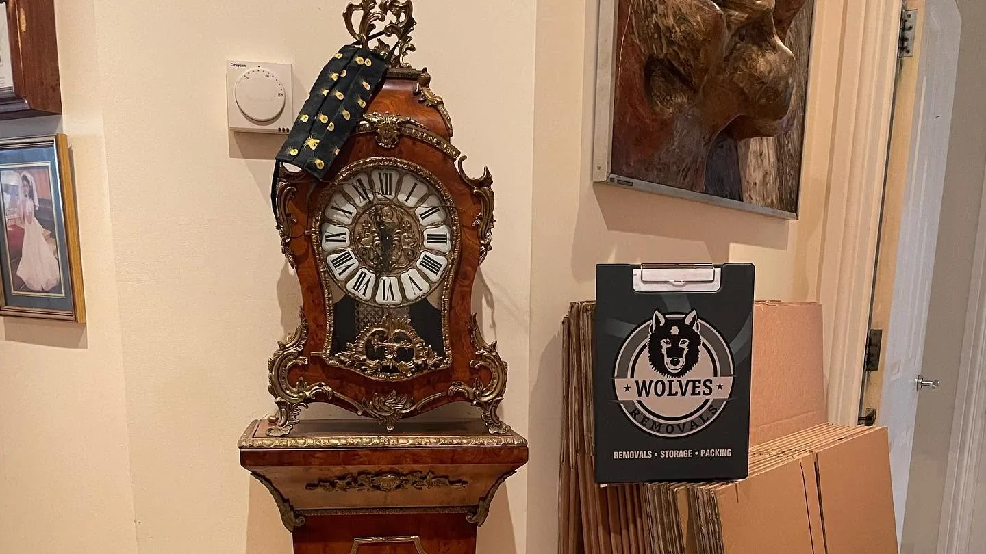 An ornate bracket clock on its pedestal beside stacked Wolves Removals packing cartons