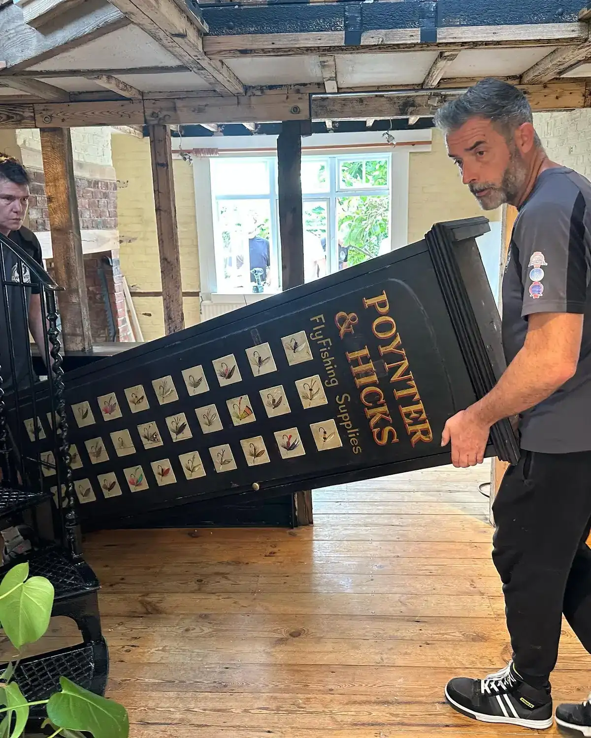 A Wolves Removals crew member carrying a large painted antique advertising board through a timber-framed room