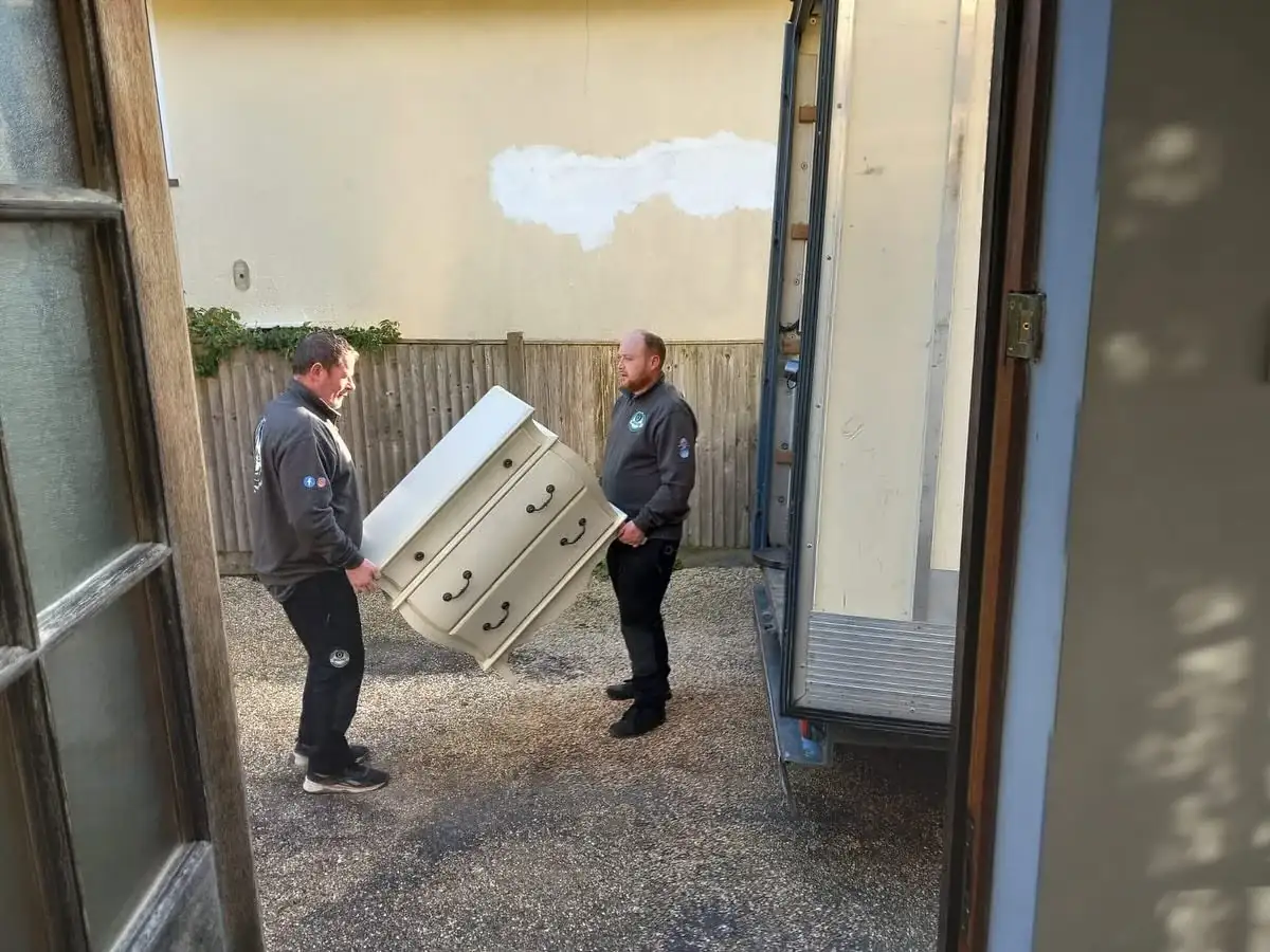 Two Wolves Removals crew carrying a painted bombe-fronted chest of drawers across gravel to a waiting van