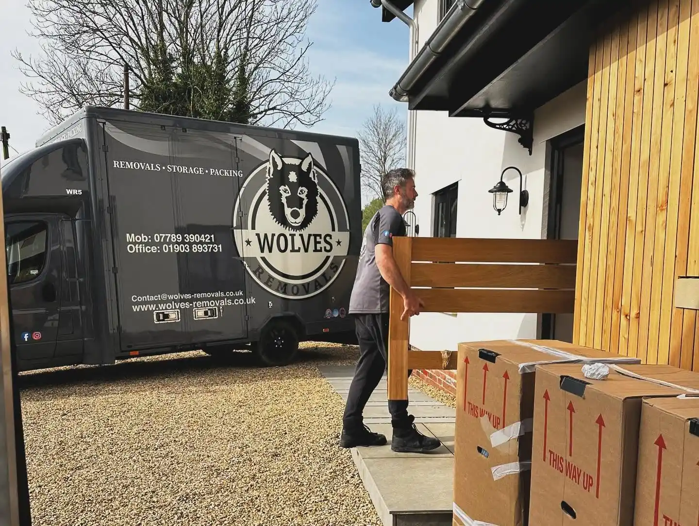 A Wolves Removals crew member carrying a timber garden bench past a stack of printed cartons at a house in Norfolk