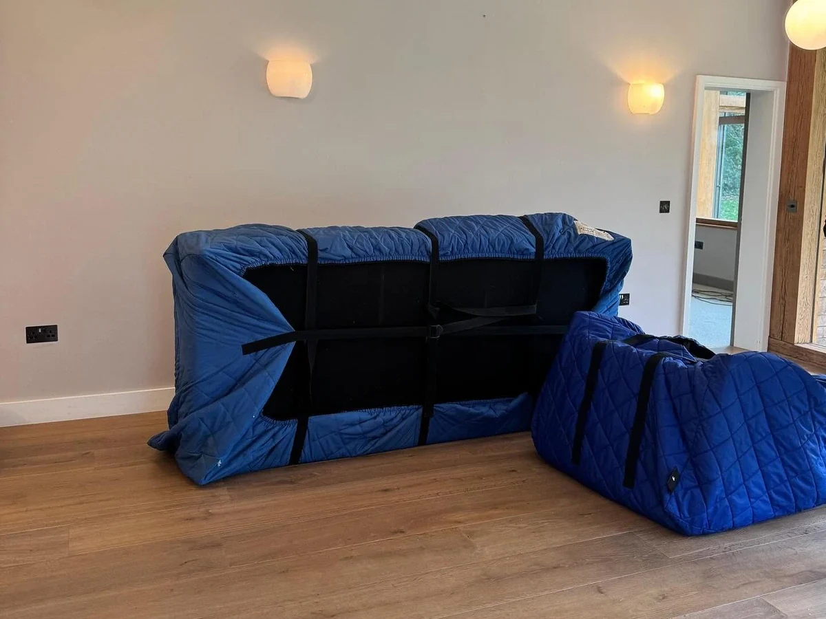 A sofa fully wrapped in quilted blue transit covers and strapped, standing in an empty room with an oak floor