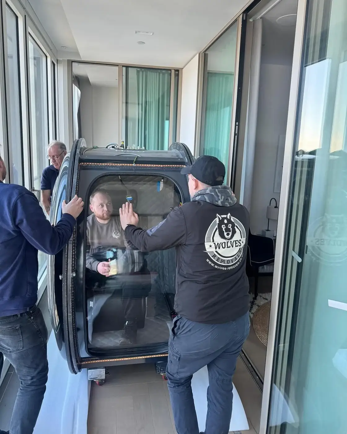 Wolves Removals crew manoeuvring a hyperbaric chamber section past floor-to-ceiling windows in a high-rise apartment