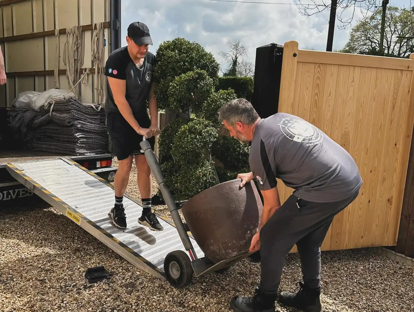 A Wolves Removals crew member steadying a large planted pot on a sack truck while another controls it from the ramp