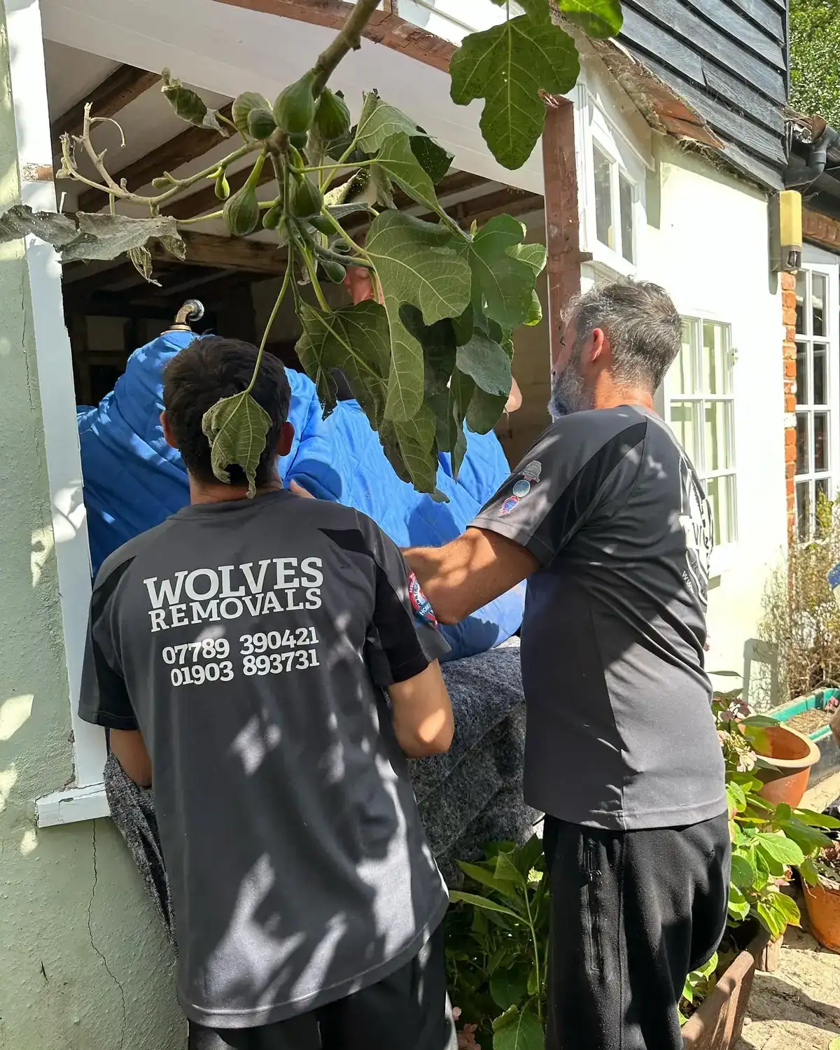Two Wolves Removals crew passing a large blue-wrapped sofa through an opening where a window unit has been removed from a period Chichester house