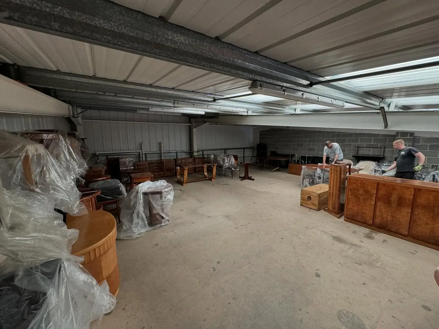 Antique furniture sheeted in clear polythene and dust covers inside a steel-framed warehouse after a removal